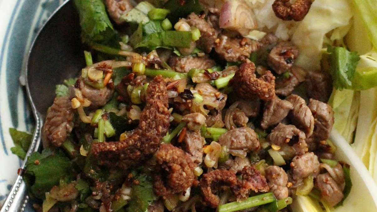 Gros plan d'un plat de Larb thaïlandais, composé de petits morceaux de viande cuite mélangés à des herbes fraîches comme la coriandre et la menthe, des échalotes tranchées, de la poudre de riz grillé et des morceaux de couenne de porc croustillante. Une cuillère en métal est plongée dans le plat, avec des feuilles de chou cru sur le côté.