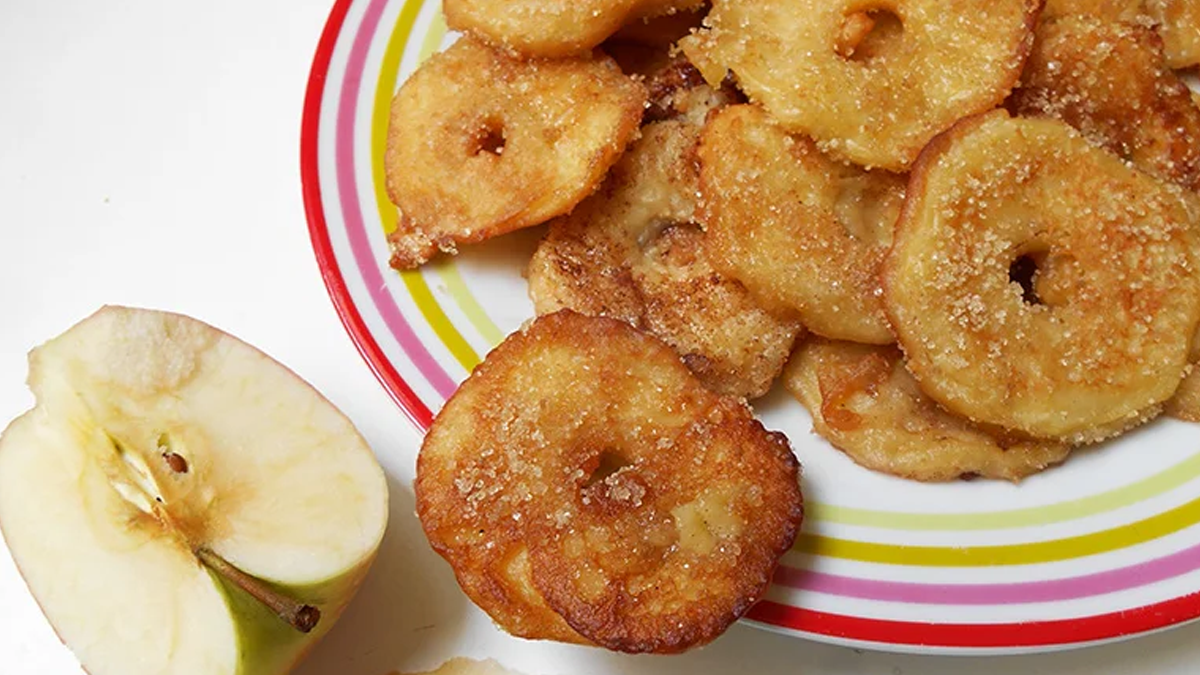 Vue de dessus d'une assiette à rayures colorées remplie de beignets aux pommes frits en forme d'anneaux, saupoudrés de sucre. Une moitié de pomme fraîche est posée à côté de l'assiette sur une surface blanche.